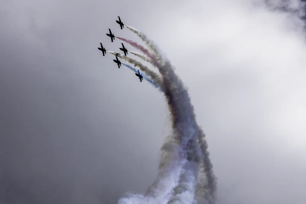Six Patriots Jet Team Aero L-39 Albatros jets climb in tight formation, trailing red, white, and blue smoke against a dramatic overcast sky during SkyFest 2026 at Fairchild Air Force Base.