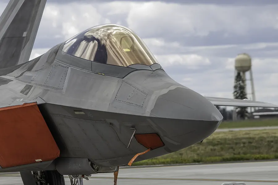 Close-up of an F-22 Raptor parked on the ramp at Fairchild Air Force Base, with its gold-tinted canopy reflecting clouds and a water tower in the background.
