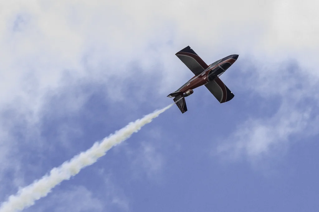 MicroJet aerobatic aircraft climbing steeply while trailing white smoke against a dramatic blue and gray cloud-filled sky.